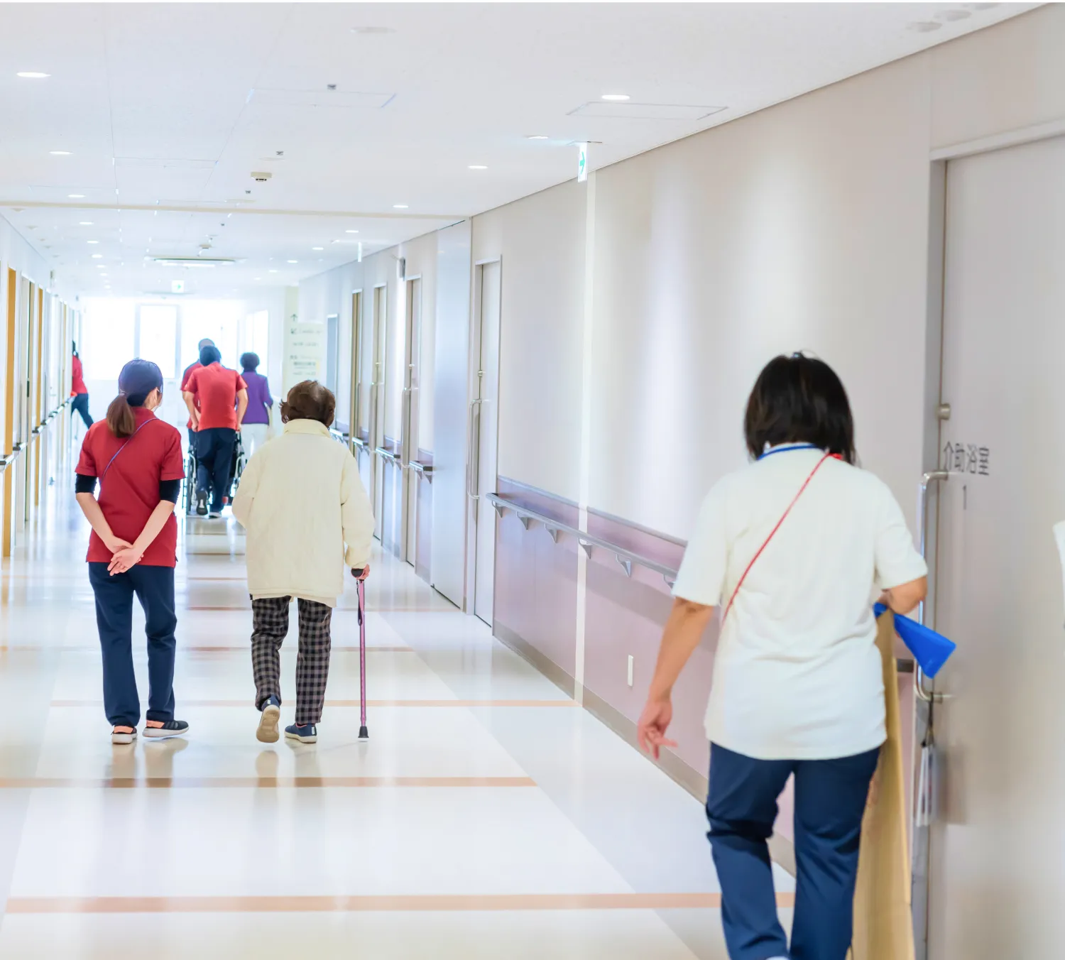 廊下で杖をついて歩いている老人とそれを見守る介護士